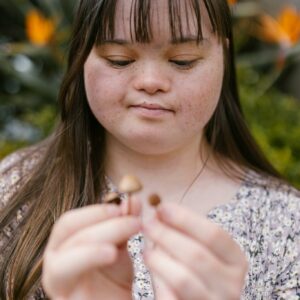 A woman with Down syndrome holds small mushrooms outdoors, surrounded by colorful flowers.