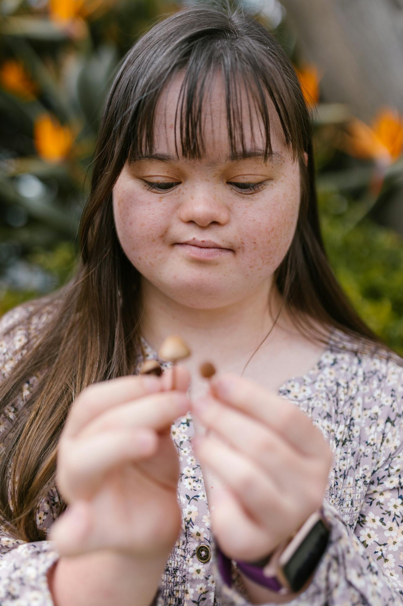 A woman with Down syndrome holds small mushrooms outdoors, surrounded by colorful flowers.