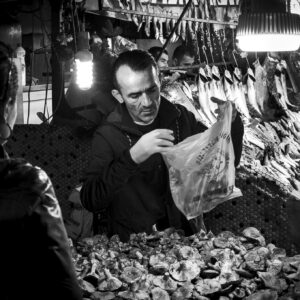 Grayscale image of a market vendor selling seafood and mushrooms at a bustling market.