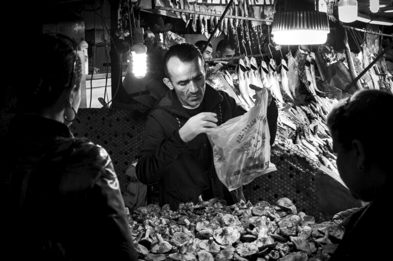 Grayscale image of a market vendor selling seafood and mushrooms at a bustling market.