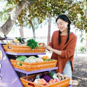 Woman in sweater selecting fresh produce at outdoor market fruit stand.